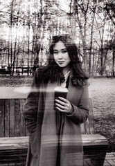 A young woman holding coffee cup stands behind a glass with reflections of trees