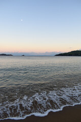 Picturesque sunset on a sandy beach in Skiathos, Greece. Gentle pastel colours glow in the sky above calm beach waves, backdropped by Greek island landscapes.