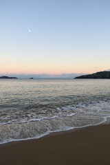 Picturesque sunset on a sandy beach in Skiathos, Greece. Gentle pastel colours glow in the sky above calm beach waves, backdropped by Greek island landscapes.