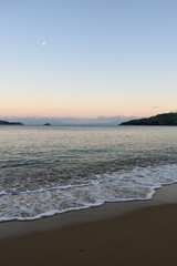 Picturesque sunset on a sandy beach in Skiathos, Greece. Gentle pastel colours glow in the sky above calm beach waves, backdropped by Greek island landscapes.