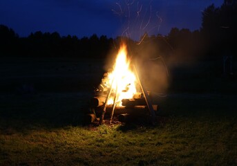 A tall pyramid-shaped bonfire made of logs blazes in the dark, sending sparks into the night sky...