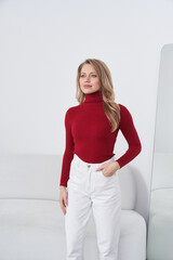 Woman wearing a red turtleneck sweater and white pants stands near a light couch, with one hand in...