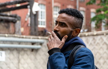 Young handsome African American man smoking cigarette in alley. Lips blowing smoke on blurred background. Portrait gangster smokes marijuana or cannabis. Portrait of a black guy smoking a cigarette.