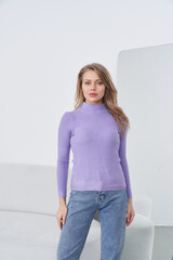 Woman with long hair wears a fitted purple sweater and light blue jeans, standing in front of a...