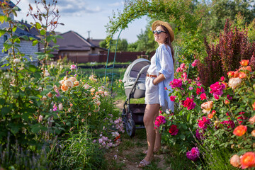 Happy young mother walking in her garden with stroller enjoying flowers. Woman gardener doing gardening chores while baby sleeps