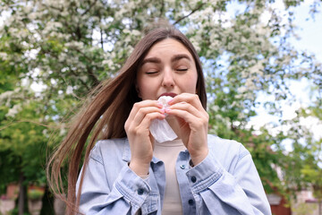 Woman with paper tissue suffering from seasonal pollen allergy near blossoming tree on spring day