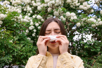 Woman with paper tissue suffering from seasonal pollen allergy near blossoming shrub on spring day