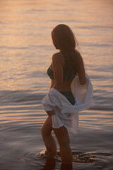 A woman in a green palm-print bikini and an unbuttoned white shirt stands in the water at dusk,...