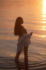 A woman in a green palm-print bikini and an unbuttoned white shirt stands in the water at dusk,...