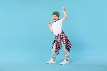 Happy little boy in baseball hat dancing on light blue background