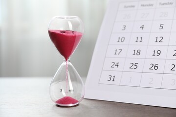 Deadline. Hourglass with flowing sand and calendar on wooden table indoors, closeup