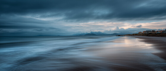 Long exposure landscape photography of beach with dramatic sky, calm water, and distant mountains at sunset