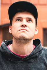A young man gazes upward with a thoughtful expression while standing in an urban area He wears a...