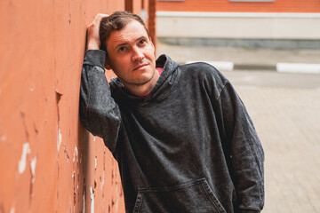 A young man in a dark hoodie leans against a weathered brick wall gazing thoughtfully toward the...