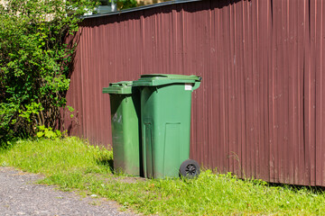 two green plastic garbage bins standing on grass by brown metal fence in sunlight outdoor waste...