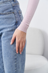 A hand rests gently on a pair of blue denim jeans. The person wears a pale pink ribbed sweater, and their fingernails are neatly manicured. A white chair is partly visible in the background