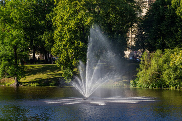 beautiful water fountain spraying in city park lake surrounded by lush green trees on sunny summer day, peaceful nature scene with reflections in water in european urban garden for relaxation