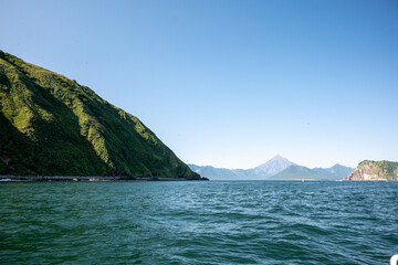 Kamchatka Peninsula. Picturesque coastal landscape with lush green mountains, clear blue sea under cloudless sky, serene view, Avachinskaya volcano is visible in the distance