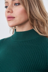 A woman with blonde hair gazes to the side, wearing a green ribbed turtleneck sweater. Her...