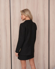 Blonde woman facing sideways, wearing a textured, black, long-sleeve blazer and short skirt. Light...