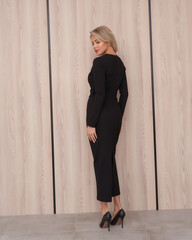 A person with blonde hair stands confidently in a fitted black dress and high heels, facing...