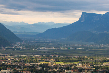 Telephoto panorama from hiking trail above Merano valley: vineyards, Adige River and layered Dolomites under patchy sunlight. Rural charm meets Alpine grandeur. South Tyrol, Italy.

