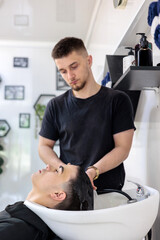 Teenager Getting Hair Washed at Modern Barbershop.  Male Barber Washing Client’s Hair at Sink in...