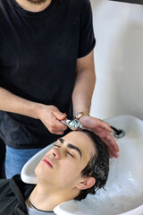 Naklejka premium Teenager Getting Hair Washed at Modern Barbershop. Male Barber Washing Client’s Hair at Sink in Hair Salon. Teenager and haircut, Men's grooming and care, Hair care for guys, Teenage beauty