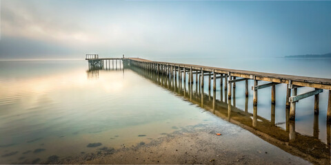 Obraz premium A wooden dock stretches over tranquil water, enveloped in soft morning mist. The peaceful atmosphere highlights the beauty of nature at dawn, inviting reflection