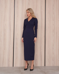 Blonde woman in elegant, long-sleeved, navy dress with V-neckline, standing on light floor with...