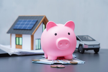 Pink piggy bank with US dollar and coins next to a miniature eco house with solar panels and an electric car model. Concept for financial goals, saving and sustainable living.
