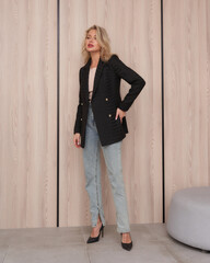 Blonde woman in a stylish black blazer, light blue jeans, and black high heels. She stands...