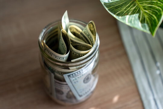 Glass jar filled with rolled US dollar bills on wooden table in natural light. Concept of personal savings, emergency fund, budgeting, money management, financial goals and frugal lifestyle at home.