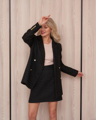 Blonde woman in a black blazer and skirt with a beige top smiles confidently, posing with one hand...
