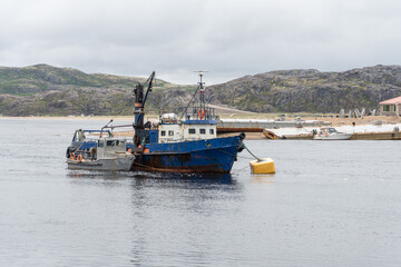 Fototapeta premium Old blue fishing vessel anchored near the shore with rugged hills in the background and cloudy sky above