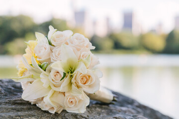Elegant bridal bouquet rests on a rock near a serene lake with city skyline in the background...