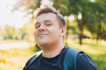 Happy 30s blonde man with backpack standing in the countryside. Traveler. Summer time. Portrait of happy young man in casual clothing standing outdoor