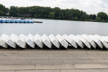 Row of white boats resting on a concrete dock beside a calm lake, surrounded by lush greenery and a...