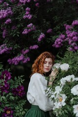 Young pretty caucasian woman in romantic outfit walking in blooming spring garden. Artistic outdoor photo of beautiful red-haired girl posing in fragrant lilac blossom.