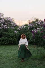 Young pretty caucasian woman in romantic outfit walking in blooming spring garden. Artistic outdoor photo of beautiful red-haired girl posing in fragrant lilac blossom.