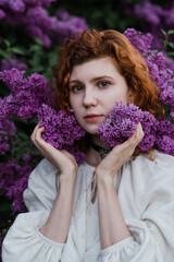 Young pretty caucasian woman in romantic outfit walking in blooming spring garden. Artistic outdoor photo of beautiful red-haired girl posing in fragrant lilac blossom.
