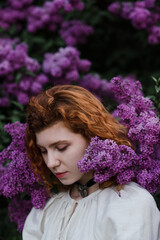 Young pretty caucasian woman in romantic outfit walking in blooming spring garden. Artistic outdoor photo of beautiful red-haired girl posing in fragrant lilac blossom.