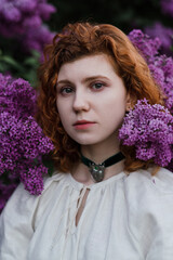 Young pretty caucasian woman in romantic outfit walking in blooming spring garden. Artistic outdoor photo of beautiful red-haired girl posing in fragrant lilac blossom.