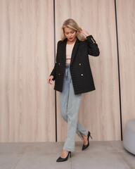 Blonde woman in stylish black blazer with gold buttons, light jeans, and black high heels stands on...