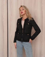 Blonde woman in a textured black jacket with gold buttons and light blue jeans poses confidently...