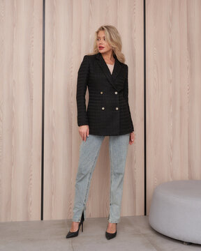 Blonde woman in black double-breasted blazer and light denim jeans, standing near a textured wooden wall. She wears black heels and has a confident expression, gazing to the side