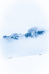 Vertical winter landscape with snowy ground, misty clouds surrounding blue-tinted mountain, bright...