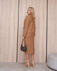Stylish woman in a tan tweed suit and nude heels, holding a small handbag. She stands against a...