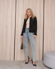 Stylish woman in high-waisted jeans, black jacket, and heels standing indoors, holding a handbag....