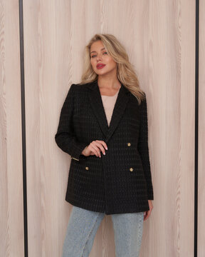 Woman in stylish black blazer with golden buttons over a light top and blue jeans, posing confidently against a vertically paneled wooden wall, with her hand on one blazer button